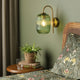 A green fluted glass wall light illuminates a bedroom with a plant, books, and floral bedding.
